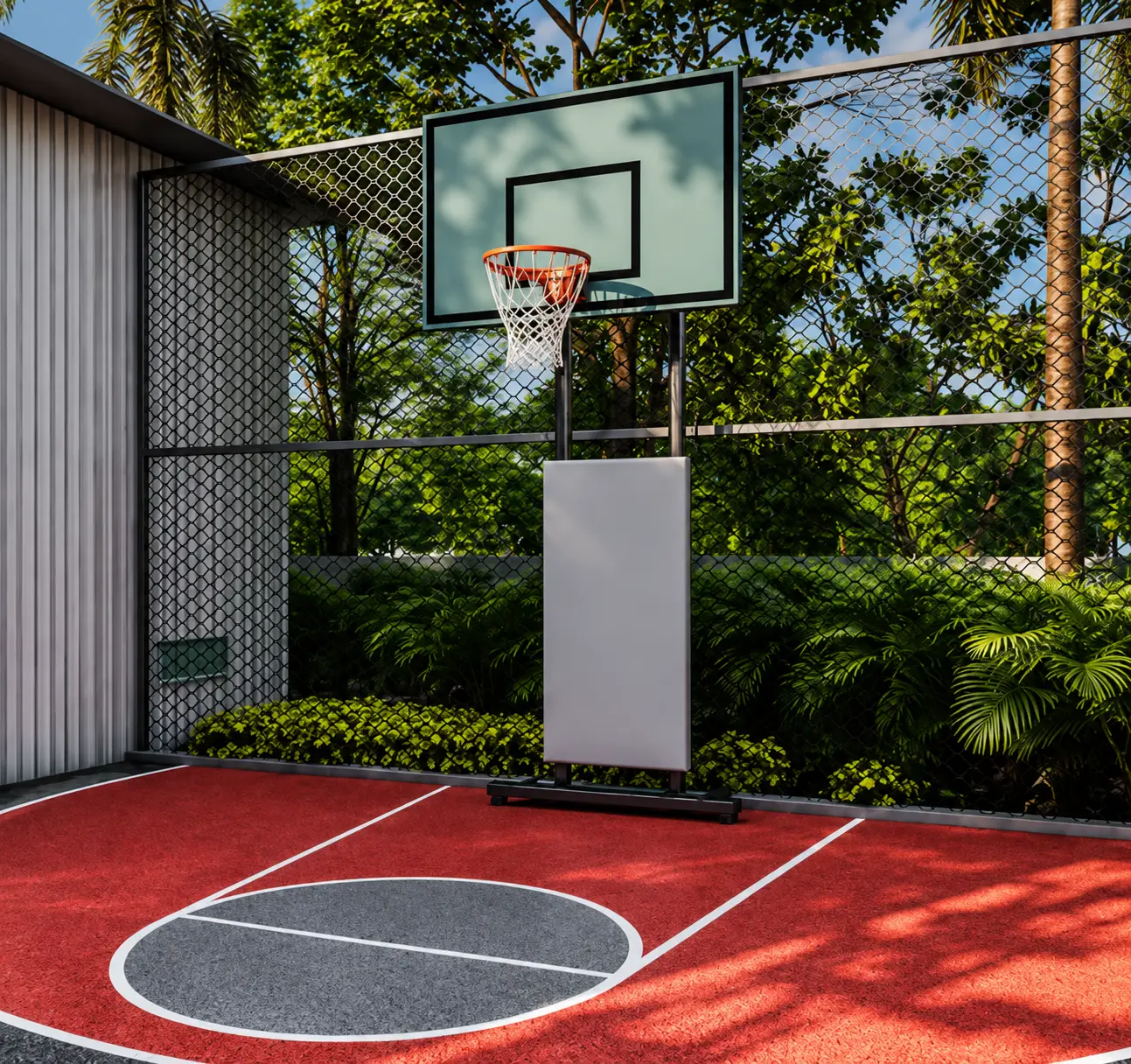 A red and grey basketball court in sunlight, a hoop on a white backboard, netted and screened by tall trees.
